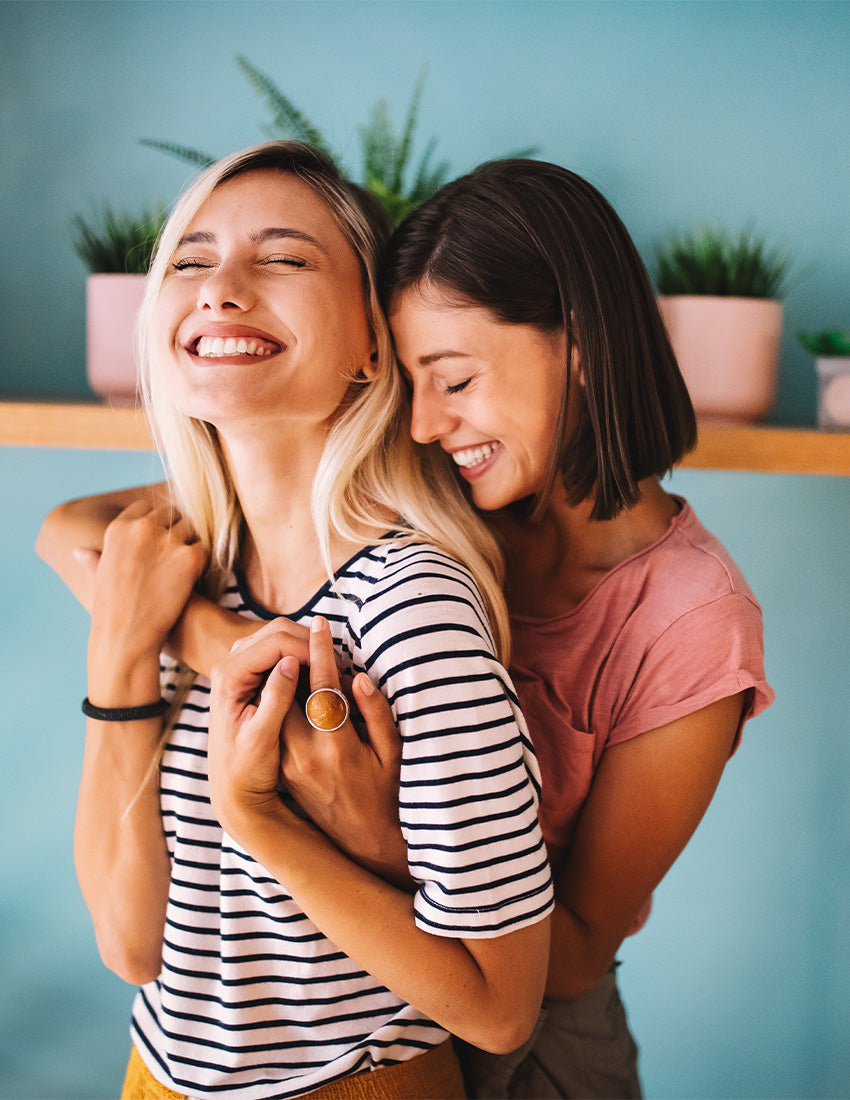 Eine Frau umarmt ihre Freundin von hinten, beide lachen und freuen sich über einen Erfolg.