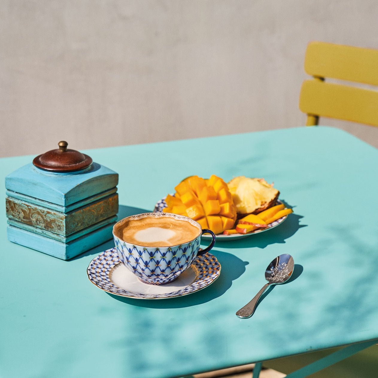 Kaffee serviert in einer blau und weiß gemusterten Tasse, eine auf einem Teller servierte aufgeschnittene Mango und eine alte Kaffeemühle stehen auf einem hellblauen Tisch. 