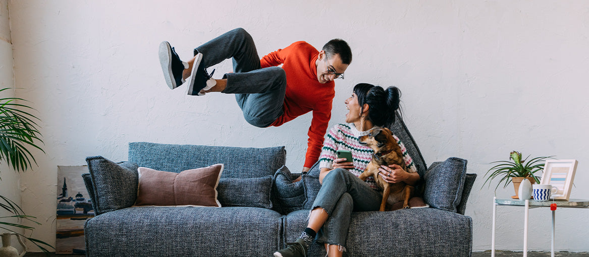 Fröhliche Szene in der ein Mann auf ein Sofa springt auf dem eine Frau mit Hund sitzt