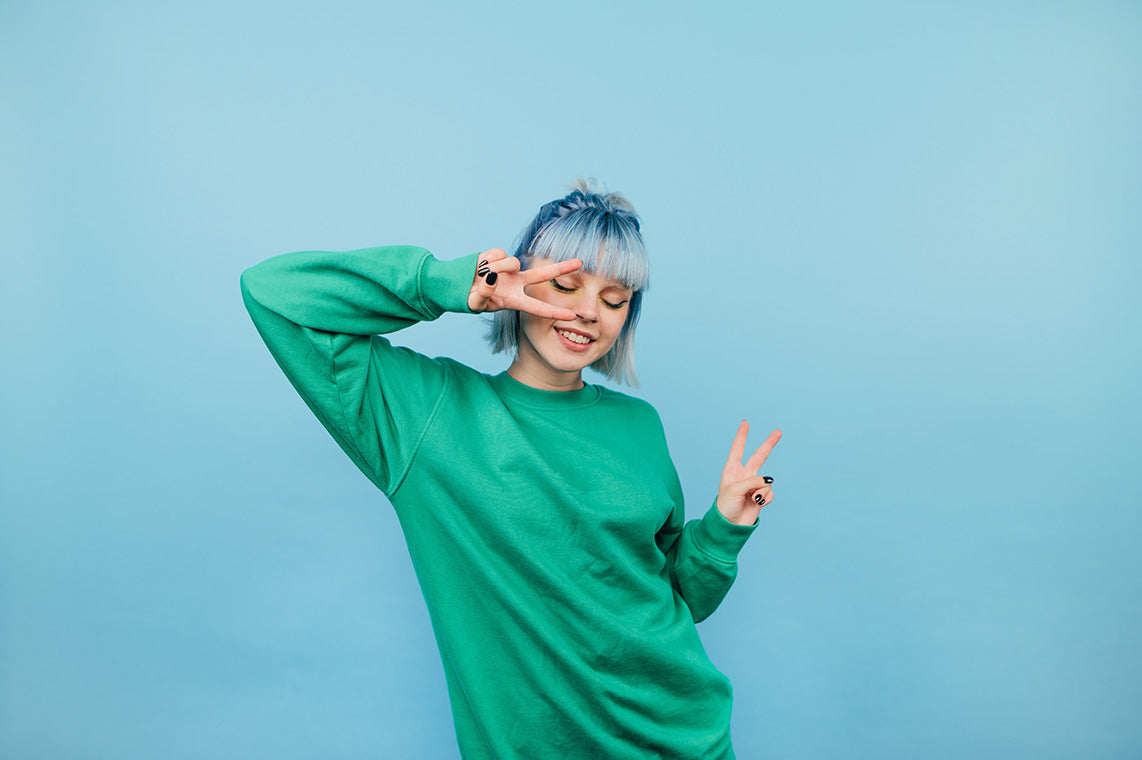 Junge Frau mit blauen Haare und grünem Pullover steht vor einer hellblauen Wand und macht mit beiden Händen ein Peace-Zeichen