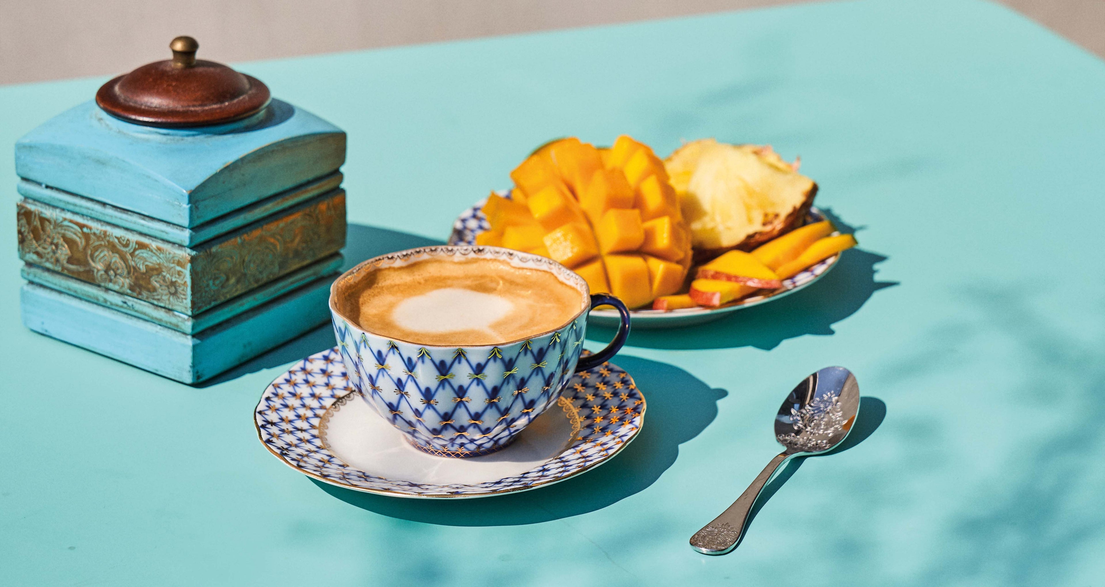 Kaffee serviert in einer blau und weiß gemusterten Tasse, eine auf einem Teller servierte aufgeschnittene Mango und eine alte Kaffeemühle stehen auf einem hellblauen Tisch. 