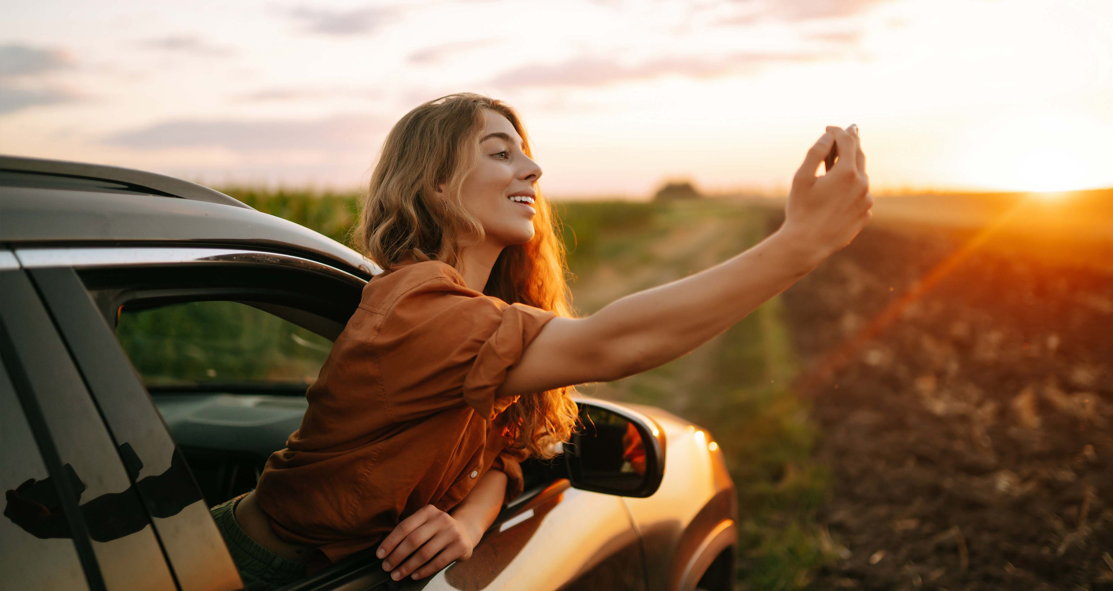 Frau lehnt sich mit Oberkörper aus dem geöffneten Beifahrerfenster eines in einem Feld geparkten Autos und macht ein Foto vom Sonnenuntergang