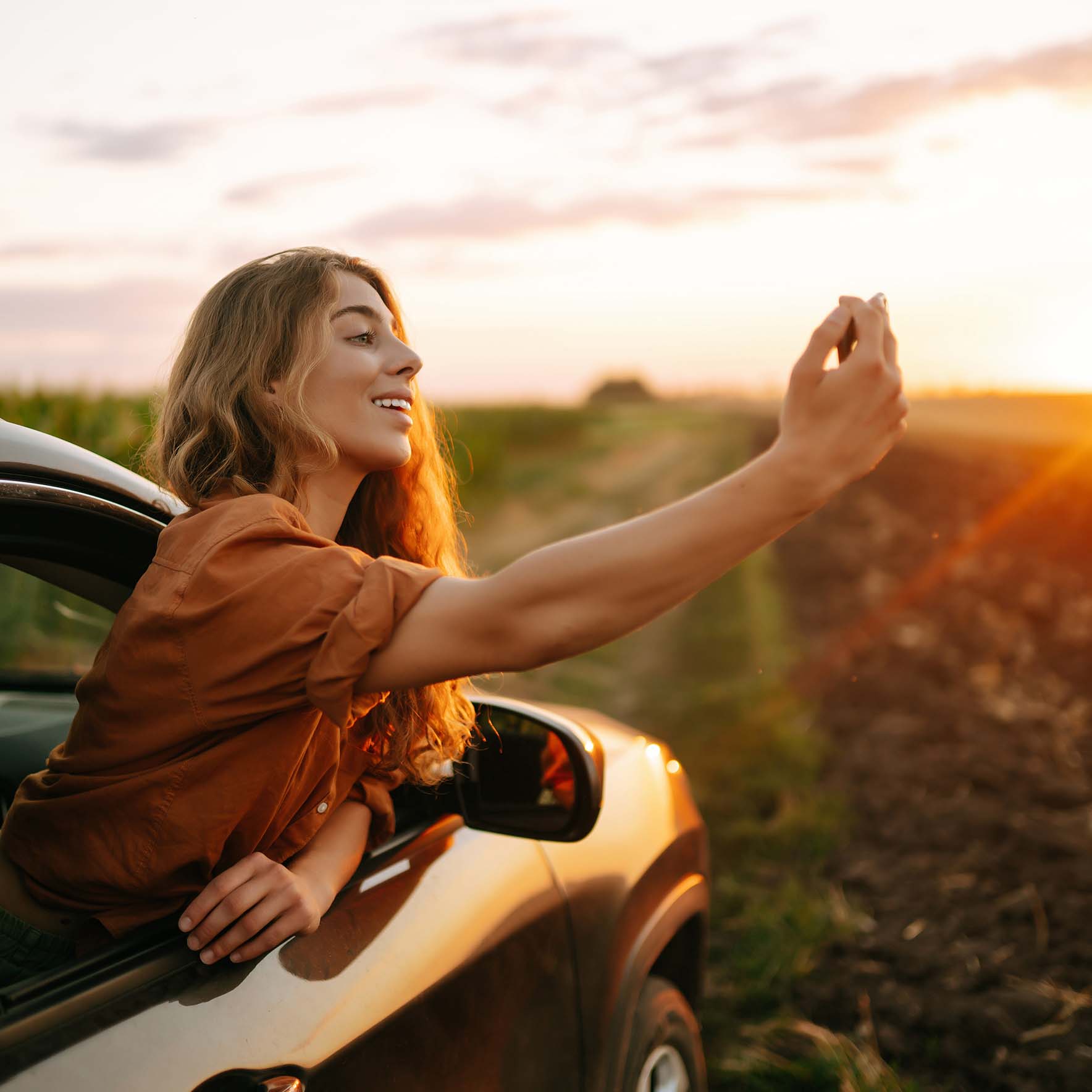 Frau lehnt sich mit Oberkörper aus dem geöffneten Beifahrerfenster eines in einem Feld geparkten Autos und macht ein Foto vom Sonnenuntergang