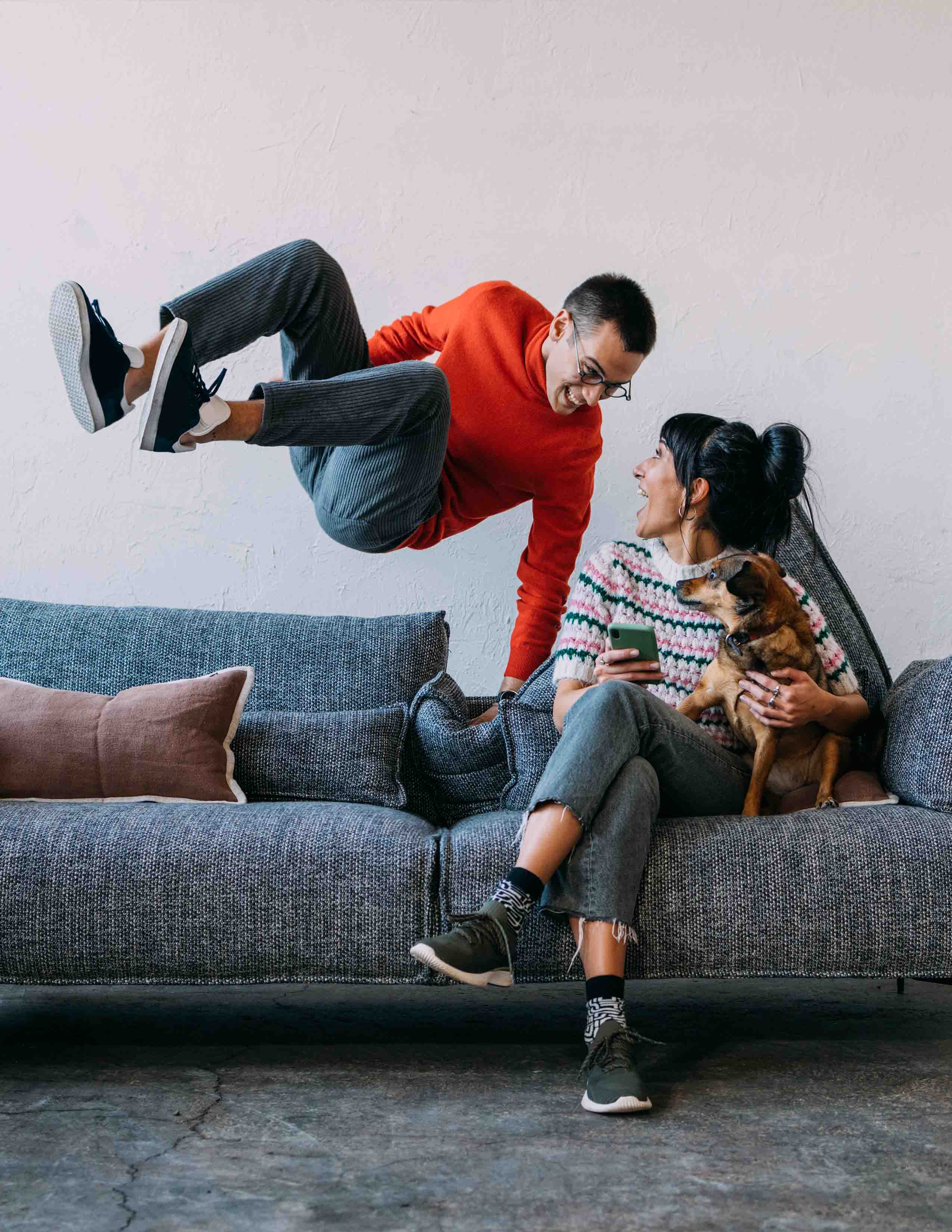 Fröhliche Szene in der ein Mann auf ein Sofa springt auf dem eine Frau mit Hund sitzt