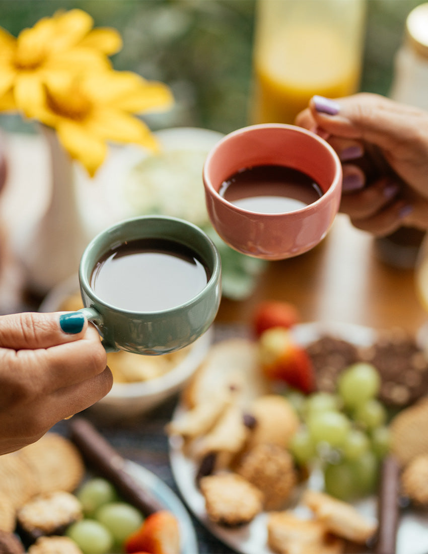 Zwei Personen, von denen nur die Hände zu sehen sind, stoßen mit Kaffee in bunten Tassen an