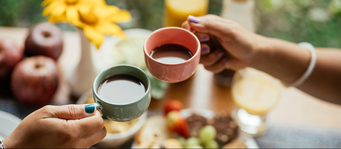 Zwei Personen, von denen nur die Hände zu sehen sind, stoßen mit Kaffee in bunten Tassen an